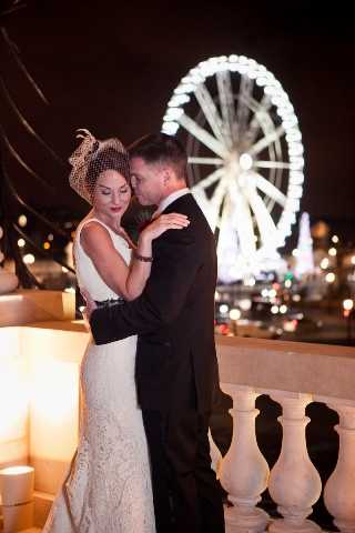 A couple shares an intimate embrace on an outdoor stone balustrade terrace at night, with the illuminated Grande Roue de Paris (Ferris wheel) and city lights visible in the background. The bride wears a white lace fitted gown with a birdcage veil and a small fascinator headpiece, while the groom is dressed in a dark suit. Candles in glass holders are visible on the balcony railing, adding warm light to the scene. This is a close-up portrait shot with a shallow depth of field, emphasizing the couple against the glowing Parisian nightscape.