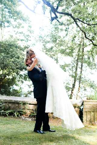 A couple portrait taken outdoors in a garden setting, where the groom, dressed in a dark navy or black suit, is lifting the bride off the ground as they embrace and kiss. The bride wears a fitted ivory lace gown with a long cathedral-length veil that cascades down as she is lifted. The setting features a manicured lawn with a low stone wall in the background and mature trees providing dappled natural backlight. The image is a full-length portrait shot with a bright, slightly overexposed background that creates a soft halo effect around the couple.