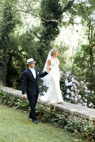 A couple portrait taken outdoors in a lush garden setting, with the bride and groom walking hand-in-hand along a low stone wall. The groom wears a black suit with a white boutonniere and a white Panama-style hat, while the bride wears a fitted white gown with a deep V-neckline and a flowing veil, carrying a small bouquet with pale pink blooms. A large flowering shrub with soft pink blossoms — likely hydrangeas — is visible behind them, adding color to the dense green foliage backdrop. The shot is a medium full-length portrait with natural daylight and a relaxed, candid feel.