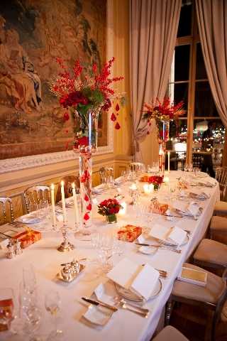 Indoor reception table setup photographed from a slightly elevated angle at one end of a long rectangular dining table covered in a white linen tablecloth. The table is set for approximately 12–16 guests with formal place settings including white folded napkins, silver flatware, and crystal glassware. Centerpieces consist of tall clear glass vases holding arrangements of red flowers and red berry branches, with hanging red crystal or glass drop ornaments adding vertical detail. Low red rose cluster arrangements and white taper candles in silver candelabras are interspersed along the table, along with small wrapped gifts or favor boxes with gold ribbon. The decor palette is bold red and white with silver accents, giving a classic and formal feel. The room features ornate wall paneling, a large decorative tapestry depicting a figurative scene, and tall windows with cream drape curtains, consistent with a Parisian hôtel particulier or chateau ballroom interior. Potential venue feature image.