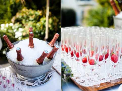 A split-image detail shot of a wedding cocktail hour beverage setup, photographed outdoors. On the left, multiple bottles of Moët & Chandon rosé champagne are chilling in a large silver ice bucket filled with crushed ice, placed on a white linen-covered table with greenery visible in the background. On the right, rows of champagne flutes are pre-filled with fresh whole strawberries and a small amount of liquid, arranged in a grid pattern on a wooden tray lined with a white doily, ready for guests to be served strawberry champagne cocktails. The setup reflects a classic French wedding reception drink service with an emphasis on presentation and detail.