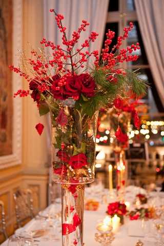 Close-up detail shot of a tall glass vase centerpiece on a white-linened reception table inside what appears to be a classic French ballroom or chateau interior. The centerpiece features deep red roses, red berry branches, pine greenery, and gold-toned dried grasses arranged in a striking vertical composition with red ribbon detail at the base of the vase. The table setting includes candles and additional lower floral arrangements with red roses visible in the background. The overall decor palette is bold red and gold with a classic winter or Christmas-inspired theme. Additional centerpieces and warm candlelight are visible in the softly blurred background, suggesting a large reception setup.