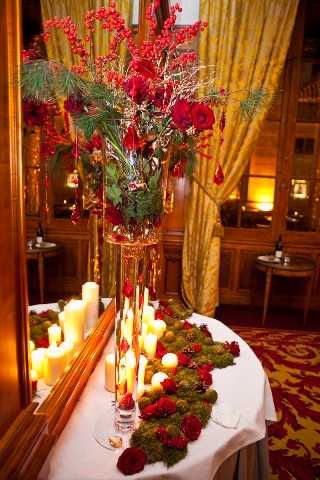 A close-up detail shot of a reception table centerpiece arrangement in an indoor ballroom setting with gold-curtained walls and warm amber uplighting. The centerpiece features a tall glass vase filled with deep red roses, red hypericum berries on bare branches, and pine greenery, with red crystal or glass drop ornaments hanging from the branches. The white-linen table is decorated with a moss runner accented by small deep red floral clusters and pine cones, flanked by varying heights of cream pillar candles in glass holders. The decor palette is deep red, green, and gold, with a formal classic style suggesting a winter or holiday-season wedding. The patterned red and gold carpet visible in the background reinforces the formal interior setting.