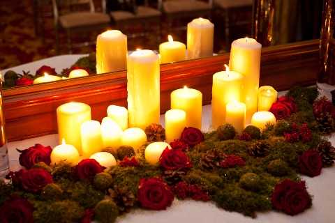 Close-up detail shot of a reception table centerpiece featuring multiple ivory and cream pillar candles of varying heights arranged on a bed of green moss, accented with deep red roses, pine cones, and small decorative spheres. A gold-framed mirror is positioned behind the arrangement, reflecting the candlelight and doubling the visual depth of the display. The overall decor palette is deep red, green, and warm gold, suggesting a winter or Christmas-season wedding theme. The warm, glowing candlelight creates a rich amber tone across the entire composition.