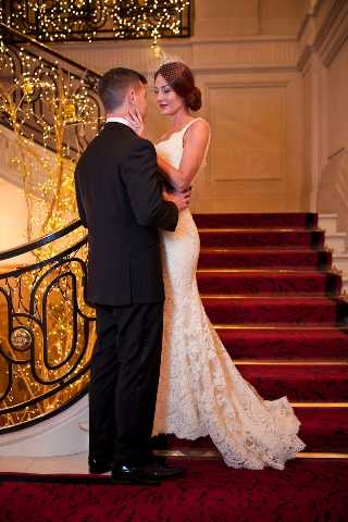 A bride and groom pose together on a grand interior staircase with deep red carpet and an ornate black wrought-iron banister wrapped in warm golden fairy lights. The bride wears a fitted ivory lace mermaid-style gown with a low back and a small birdcage veil, while the groom is dressed in a classic black tuxedo. The couple faces each other in a close embrace, with the bride standing one step above the groom. The portrait-style shot captures the full length of both figures against the richly decorated staircase backdrop, suggesting a formal ballroom or hotel venue. Potential venue feature image.