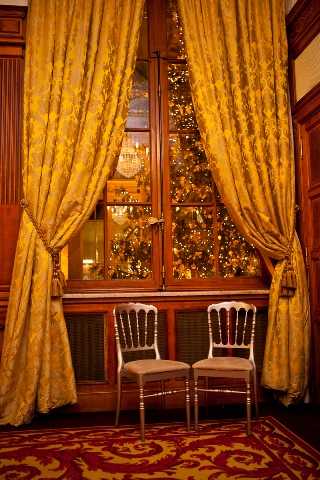 Detail shot of an interior venue space featuring two gold Chiavari chairs positioned side by side in front of a tall wooden-framed window flanked by floor-length gold damask curtains with tassel tiebacks. Through the window, a warmly lit Christmas tree decorated with golden fairy lights is visible, suggesting a winter or holiday-season wedding. The flooring is covered in a deep red and gold patterned carpet, and the wood-paneled walls contribute to a classic, formal interior aesthetic. The overall decor palette is rich gold and burgundy. Potential venue feature image.