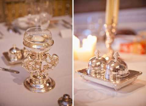 Close-up detail shots of ornate silver tableware placed on white linen-covered reception tables. The left image shows an elaborate silver candelabra or compote stand with decorative figurines at its base holding a glass bowl, accompanied by additional small silver condiment pieces. The right image shows a polished silver butter dish with a domed lid on a rectangular tray, with lit ivory taper candles softly blurred in the background. The decor reflects a classic, formal table styling with warm candlelight and a silver and white color palette. Side-by-side detail composition.