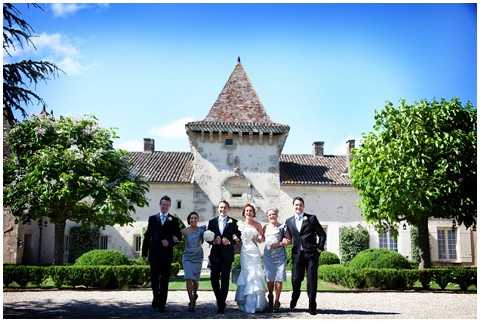 A bridal party of six walks toward the camera in a wide outdoor shot, arms linked and smiling, in front of a French chateau featuring a distinctive pointed stone tower with terracotta roof tiles and arched entryway. The bride wears a full-length white ball gown, while the two bridesmaids wear knee-length silver-grey dresses; the three groomsmen are dressed in dark suits. The group is positioned on a gravel courtyard flanked by trimmed hedges and mature trees. Potential venue feature image.