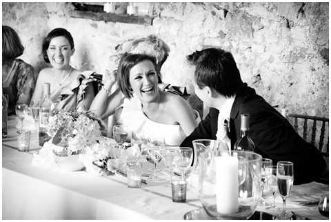 A black-and-white image capturing a candid moment during the wedding reception at what appears to be an indoor venue with exposed stone walls. The bride, wearing a strapless white dress and a decorative hair piece, is laughing openly while seated at the head table alongside the groom in a dark suit jacket. A female guest visible in the background is also laughing and appears to be holding a camera. The table is set with glassware, candles, wine bottles, and low floral centerpieces. The image is a mid-range shot with high contrast black-and-white tones that emphasize the joyful expressions of those pictured.