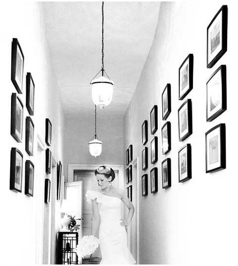 Black-and-white portrait of a bride standing in a narrow interior hallway, looking downward with a relaxed pose. She wears a strapless or one-shoulder white fitted gown and holds a round bouquet of what appear to be peonies or ranunculus, rendered in bright white tones against the darker dress fabric. Her hair is styled in an updo with a small decorative piece. The hallway features two pendant lantern-style light fixtures hanging from the ceiling and both walls are densely lined with framed artwork or photographs in dark frames, creating a gallery-wall effect. The image is a full-length portrait shot taken from the far end of the hallway, using the architectural lines to draw the eye toward the bride.