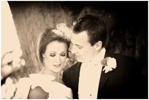 A sepia-toned close-up portrait of a bride and groom standing together outdoors, both looking downward. The bride wears an off-shoulder dress and a lace or tulle hair piece, with styled updo hair and defined eye makeup; she holds what appears to be a bouquet of light-colored flowers. The groom wears a dark tuxedo jacket with a white shirt and a light-colored boutonniere on his lapel. The warm sepia tone gives the image soft contrast with lighter areas around the subjects and a darker background of textured tree bark.