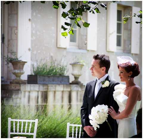 A couple stands together outdoors in front of a French chateau or manor house facade with white shuttered windows and stone urns. The groom wears a dark charcoal suit with a white boutonniere and dark tie, while the bride wears a strapless white dress with a large bow or ruffle detail at the hip and carries a round bouquet of white peonies or ranunculus; she also wears a small white birdcage veil and has auburn upswept hair. The setting suggests an outdoor ceremony or portrait moment, with white chiavari chairs visible in the foreground. The shot is a medium portrait composition capturing both figures from roughly the waist up, with a classic, clean white and dark contrast styling theme.
