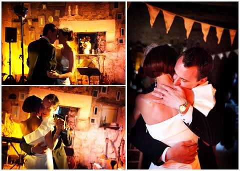 A collage of three reception photos taken in a rustic indoor venue with warm amber lighting and exposed stone walls. The top-left image shows the bride and groom sharing a close moment on the dance floor, with stage lighting and a fireplace visible in the background. The bottom-left image captures the bride photographing something with a camera, illuminated by a warm spotlight. The large right image shows the couple in a close embrace, with the bride wearing a strapless white gown with a large bow detail at the back and the groom in a dark suit with a gold watch; orange and natural-toned fabric bunting is visible overhead. The overall styling is rustic and relaxed, shot in a warm, low-light reception atmosphere.