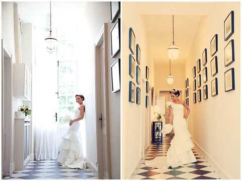 A diptych of two bridal portraits taken in the same interior hallway of what appears to be a French chateau or manor house, featuring a bride in a strapless white mermaid-style gown with a ruffled train and a small floral headpiece, holding a white bouquet. The hallway has a distinctive black-and-white diamond-patterned tile floor, cream walls lined with navy blue-framed artwork or mirrors arranged in a gallery-wall style, and a glass pendant light fixture overhead. The left image is cooler-toned and shows the bride in profile near a bright windowed door at the end of the corridor, while the right image has a warmer yellow tone with the bride facing slightly toward the camera in a three-quarter pose. Both shots are full-length portraits capturing the architectural details of the venue interior. Potential venue feature image.