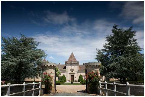 Wide-angle exterior shot of a French château wedding venue, viewed from the entrance gate along a gravel driveway. The stone château features a central conical tower, crenellated walls, and a formal arched entrance doorway, flanked by large mature cedar trees and climbing roses in red and pink tones. White wooden gate posts frame the foreground, leading the eye toward the building. No people are visible in the frame. Potential venue feature image.