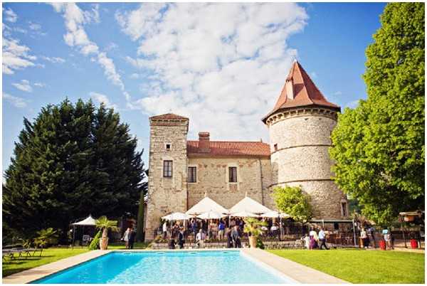 A wide-angle outdoor shot of a wedding cocktail hour taking place on the grounds of a stone French chateau featuring a distinctive round tower with a terracotta-tiled conical roof. A large group of approximately 30-40 guests mingles on the terrace directly in front of the chateau, shaded by several white market umbrellas. In the foreground, a rectangular swimming pool with bright blue water occupies the lower portion of the frame. The overall aesthetic is classic French countryside. Potential venue feature image.