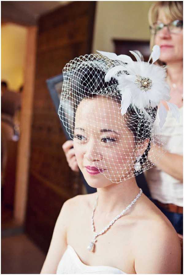 A getting-ready portrait of a bride being assisted by a stylist or family member in an indoor setting with warm, natural light. The bride wears a white strapless dress and a delicate pearl drop necklace, with her hair styled beneath a white birdcage veil topped with a feather fascinator featuring white and black feathers and a gold brooch centerpiece. Her makeup includes a berry-pink lip. The stylist, visible in the background, is adjusting the veil. The composition is a close-up portrait with a shallow depth of field, keeping the background softly blurred.