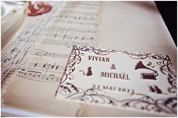 Close-up detail shot of wedding stationery for Vivian and Michaël, dated 11 Mai 2013. The invitation or program features a vintage-inspired design printed in dark brown ink on cream paper, incorporating decorative flourishes, silhouette illustrations of a cat, a gramophone, and other small motifs. Layered beneath is a sheet of printed musical notation, adding to the vintage, music-themed styling. A lace trim is visible along the edge, and a partial red wax seal can be seen in the upper left corner.