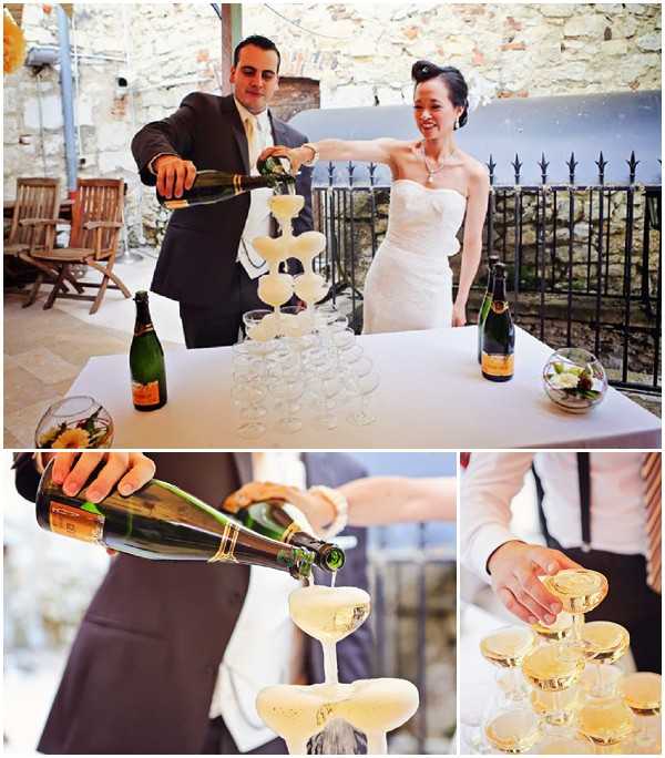 A three-image collage documenting a champagne tower pouring moment during a wedding reception. In the main top image, the groom, wearing a dark suit with a cream tie, pours champagne from a green bottle over a tiered tower of coupe glasses on a white-clothed table, while the bride, in a strapless ivory gown with her hair up, assists and laughs. Two Veuve Clicquot bottles and small floral centerpiece bowls are visible on the table, set on a covered outdoor terrace with stone walls and a black iron railing in the background. The bottom-left detail shot shows champagne cascading from a bottle into the top coupe glass in close-up, and the bottom-right close-up shows hands steadying the stacked coupe glasses, which have a warm gold tone from the champagne filling them. The overall decor palette is classic and minimal, with white linens and clear glassware.
