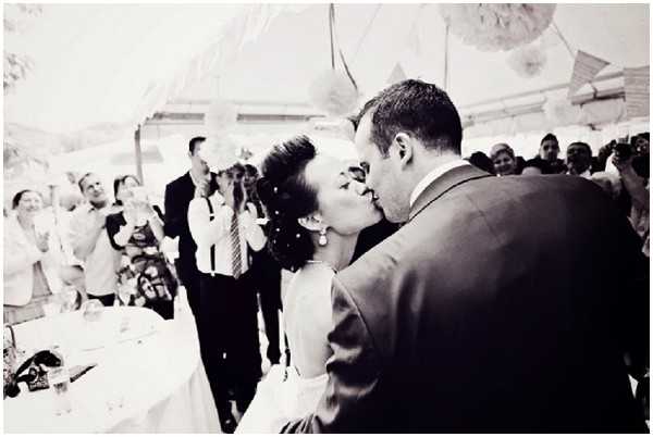 Black-and-white close-up portrait of a bride and groom sharing a kiss, shot from behind the groom's right shoulder with the couple centered in the frame. The bride has her hair styled up and wears drop earrings and a strapless or sleeveless dress, while the groom wears a dark suit jacket. The setting appears to be an outdoor or semi-outdoor reception under a white tent or marquee, with decorative paper lanterns or pompoms visible overhead. A crowd of approximately 15–20 guests is visible in the background, many smiling, clapping, or photographing the moment, with a reception table partially visible to the left.