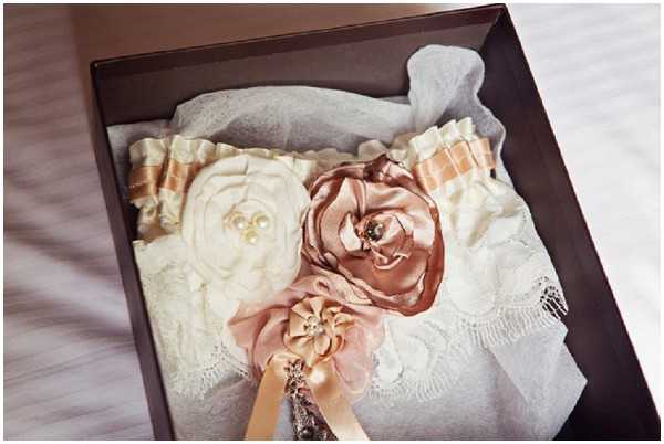 Close-up detail shot of a bridal accessory set displayed inside a dark brown gift box. The arrangement includes a white lace garter embellished with a large ivory fabric rosette accented with pearl beading, a mauve-pink satin rosette, and a rose-gold or copper-toned satin rosette, along with a striped champagne and gold ribbon. A smaller blush pink fabric flower with a ribbon tail is also visible in the lower portion of the box. The items rest on white lace and tissue paper, suggesting a getting-ready or flat-lay preparation scene. The styling is romantic and vintage-inspired, with a soft, warm color palette of ivory, blush, and rose gold.