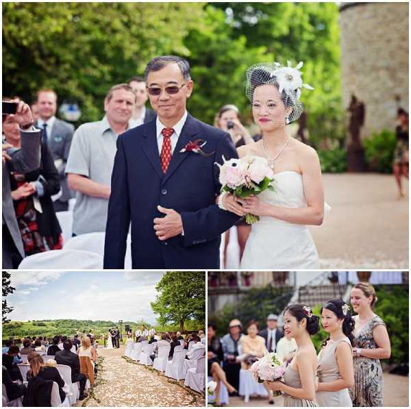 A collage of three images from an outdoor wedding ceremony at what appears to be a French chateau or vineyard estate. The top image shows the bride walking down the aisle escorted by a man in a navy suit with a red tie and boutonniere; the bride wears a strapless ivory gown, a birdcage veil with a white floral fascinator, a pearl necklace, and carries a bouquet of blush pink and white blooms including peonies. The bottom-left image is a wide shot of the outdoor ceremony aisle lined with white chairs and flower petal scattered on the ground, with rolling vineyard hills visible in the background under a bright sky. The bottom-right image shows two bridesmaids in silver-grey dresses, one holding a bouquet of blush pink flowers, smiling during the processional, with seated guests visible behind them. The overall styling is classic with a vintage touch, shot in natural daylight. Potential venue feature image.