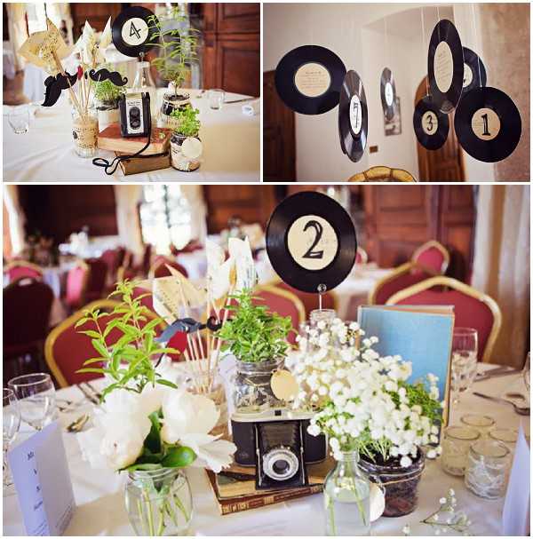 A three-image collage showing wedding reception table decor with a vintage music theme, set in an indoor venue with dark wood paneling and red upholstered banquet chairs. The centerpieces are built around stacks of vintage hardcover books topped with antique film cameras, surrounded by small potted green herbs, white peonies, white baby's breath in glass jars, and novelty photo booth props on sticks including paper mustaches and fans. Table numbers are displayed on vinyl records mounted on stands, with cream-colored number labels at the center of each record. The top right image shows additional vinyl records hung as a garland displaying table numbers 1, 3, and others. The color palette across the decor is black, white, cream, and green, with the vintage cameras and worn book spines adding warm brown tones. The overall styling theme is retro-vintage with a music and photography motif. Detail and wide reception shots are both represented in the collage.