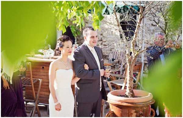 A bride and groom stand together outdoors during what appears to be a cocktail hour or reception gathering. The bride wears a strapless white fitted gown with a statement necklace and bracelet, her dark hair styled up, while the groom wears a dark charcoal suit with a white dress shirt and tie, holding a champagne flute. The setting is a rustic courtyard or terrace with stone walls, wooden bistro chairs and tables, and a large terracotta planter holding a bare-branched tree. Several guests are visible in the background, seated and mingling. The overall styling is classic and understated, with a candid portrait composition framed by green foliage in the foreground.