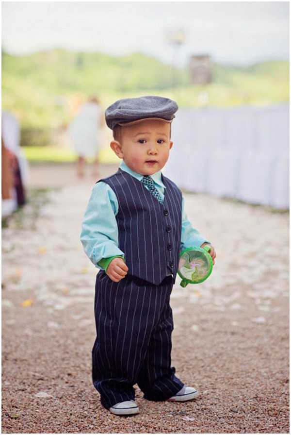 A close-up portrait of a toddler boy standing on a gravel path at an outdoor wedding venue. He is dressed in a navy blue pinstripe vest and matching trousers, a mint green button-up shirt, a small patterned bow tie, a grey flat cap, and white low-top canvas sneakers. He is holding a small green plastic toy or sippy cup in one hand. In the blurred background, a figure in a white dress is visible along with what appears to be a draped white tent or table covering, suggesting a reception setting.