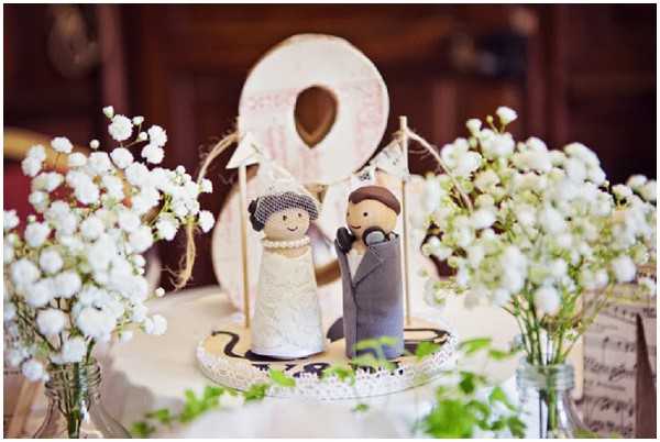 Close-up detail shot of a wedding cake topper featuring two handmade wooden peg dolls dressed as a bride and groom — the bride in a white lace dress with a birdcage veil and the groom in a grey suit — displayed on a small round lace-trimmed base. Surrounding the topper are several small glass jar vases filled with white baby's breath, and a decorative wooden letter 'O' with a heart cutout and distressed white paint is positioned behind the figures. Sheet music cards are visible in the background, contributing to a rustic, whimsical decor theme with a white and natural wood color palette.