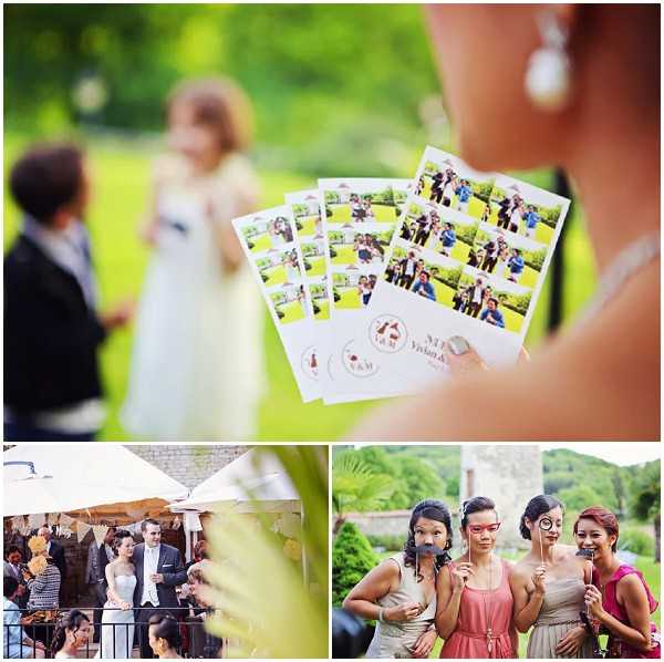 A collage of three images from an outdoor wedding reception. The main top image is a close-up detail shot of a guest's hand holding four photo booth strip printouts featuring wedding guests posing in various groups, each strip branded with a custom stamp reading 'Vivian & M' with a couple silhouette logo; in the soft-focus background, a bride in a white dress and groom in a dark suit are visible conversing with guests on a green lawn. Bottom left shows a wide shot of the couple — bride in a strapless white gown, groom in a dark suit — standing on a terrace or balcony of a stone building decorated with bunting, surrounded by seated and standing guests during what appears to be a cocktail hour or reception. Bottom right features a portrait of three bridesmaids posing playfully with photo booth props including mustaches and glasses on sticks; they wear a mix of coral/pink and champagne-taupe dresses, with a stone building and lush greenery visible in the background.