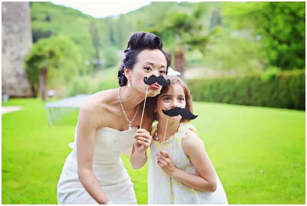 A bride and a young girl pose together outdoors on a lawn, both holding black paper moustache props on sticks up to their faces in a playful moment, likely during a cocktail hour or reception. The bride wears a strapless ivory gown with an upswept hairstyle and a pearl necklace, while the young girl wears a light yellow floral dress with a small hair accessory. The setting appears to be the grounds of a chateau or estate, with a manicured lawn and wooded hillside visible in the background. The shot is a medium portrait with a shallow depth of field, giving a candid, lighthearted feel.
