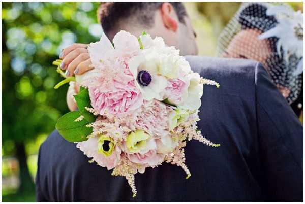 Close-up detail shot of a bridal bouquet held by the bride as she embraces the groom outdoors, with soft green foliage visible in the background. The bouquet features large blush pink peonies, a white anemone with a deep purple center, pale yellow lisianthus, and delicate blush astilbe, accented with green leaves. The groom is wearing a dark charcoal suit, and the bride is partially visible wearing a black birdcage veil with white feather accents. The composition keeps the couple slightly out of focus, drawing attention to the bouquet in the foreground.