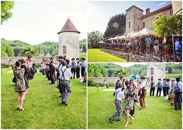 A collage of three images from an outdoor wedding cocktail hour or reception at a French chateau or manor property. The grounds feature a round stone tower with a terracotta-tiled conical roof, well-maintained green lawns, and stone walls. Approximately 20-30 guests are visible across the images, many wearing traditional French folk attire including black wide-brimmed hats and suspenders, suggesting a regional folk dance or farandole is taking place on the lawn. In the top-right image, guests line a terrace railing overlooking the grounds, with white market umbrellas providing shade. The overall styling is rustic and festive, with a folk/regional cultural theme. Wide shots capture the activity and venue grounds together. Potential venue feature image.