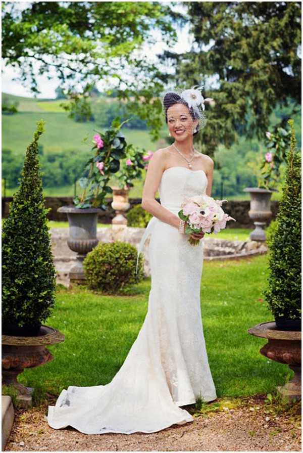 A bridal portrait taken outdoors in a formal garden setting, featuring a single bride standing between two trimmed conical topiary trees in stone urns. She wears a strapless ivory fitted lace gown with a train, paired with a pearl necklace and a white birdcage veil fascinator accented with a feather and flower. She holds a small bouquet of blush pink and white anemones with pearl-wrapped stems. The background includes additional stone urn planters with pink climbing roses and rolling green hills in the distance. The styling has a classic vintage feel. Medium full-length portrait shot.