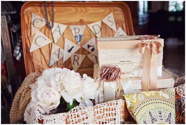 A close-up detail shot of a rustic-boho wedding card and gift display station. A wicker basket trimmed with cream crochet lace holds a cluster of white peonies, and a burlap-and-pennant bunting spelling 'CARDS' is strung across a wooden tray in the background. Nearby are stacked keepsake boxes wrapped in cream lace and tied with a peach satin ribbon bow, a small pouch tied with a mauve fabric flower and pearl strand, and a yellow and green patterned fabric item in the foreground. The overall decor palette is ivory, cream, peach, and muted gold with a vintage-rustic styling theme.