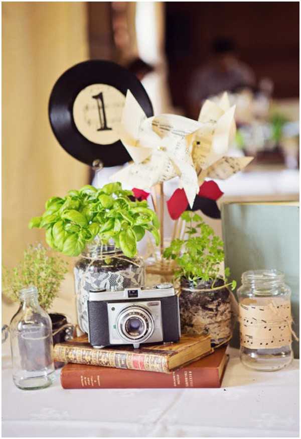 Close-up detail shot of a wedding reception table centerpiece with a vintage and eclectic styling theme. The centerpiece features a black vinyl record used as a table number marker displaying the number '1', alongside paper pinwheels made from vintage sheet music pages and red paper. Mason jars wrapped in lace and burlap ribbon hold potted fresh herbs including basil and thyme, while an empty glass bottle sits to the left and a jar decorated with sheet music paper sits to the right. A vintage film camera rests on top of a stack of antique hardcover books at the center of the arrangement. The overall decor palette combines cream, black, red, and natural green tones with a retro, DIY-crafted aesthetic.