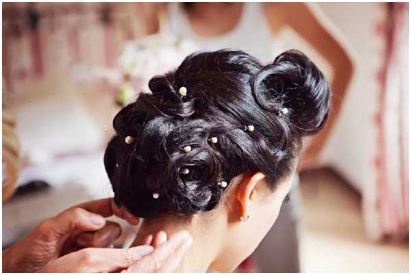 A close-up detail shot of a bride's updo hairstyle being finished by a stylist during the getting-ready phase. The bride has dark brown-black hair styled into an elaborate pinned updo with curled and twisted sections, decorated with small pearl-tipped hairpins scattered throughout. A pair of hands, presumably the hairstylist's, are visible at the nape of the neck making final adjustments. The background is softly blurred, showing what appears to be an indoor preparation room with light-colored walls and a mirror reflection.