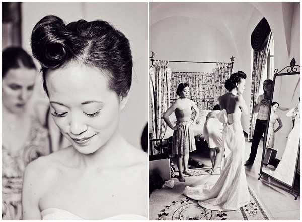A black-and-white diptych capturing a bride getting ready indoors, likely in a chateau or villa room with arched windows and ornate architectural details. The left panel is a close-up portrait of the bride looking downward, showcasing her vintage-style updo with large rolled curls pinned at the crown and her strapless gown. The right panel is a wider shot showing the bride in her strapless white gown with a corseted back and train, standing before a full-length mirror while an assistant or bridesmaid helps fasten the dress; a second attendant in a knee-length dress stands nearby. The black-and-white tones are soft with moderate contrast, giving the images a classic editorial feel.