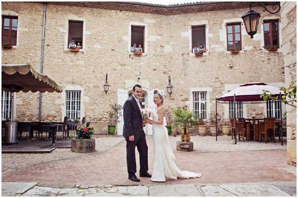 A bride and groom pose together in a stone courtyard in front of a large rustic French limestone building, likely a chateau or domaine. The bride wears a slim-cut ivory gown with a small white headpiece and carries a compact white bouquet, while the groom wears a dark navy suit with a white shirt and tie. The courtyard features terracotta pots with plants, wrought-iron wall lanterns, and outdoor dining furniture visible under awnings on both sides. Guests can be seen leaning out of the upper-floor windows watching the couple below. This is a wide portrait shot with the venue building as a prominent backdrop. Potential venue feature image.