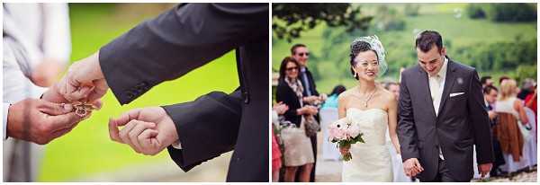 A two-panel image from an outdoor wedding ceremony. The left panel is a close-up detail shot of two men's hands exchanging or handling wedding rings, with the groom wearing a dark suit jacket and white dress shirt visible. The right panel shows the bride and groom walking hand-in-hand as a couple after the ceremony, surrounded by guests on both sides in an outdoor garden or countryside setting with green rolling hills in the background. The bride wears a strapless ivory dress with a small birdcage fascinator in her hair and carries a small bouquet of blush pink roses, while the groom wears a charcoal grey suit with a white pocket square. Both are smiling and looking downward as guests look on, with approximately 10-15 guests visible in the background.