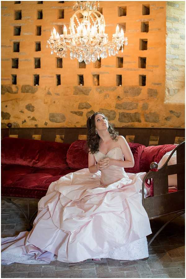 A bridal portrait of a single bride seated on a dark wood and deep red velvet settee or antique bench, set against a rustic stone wall with a decorative grid-cut pattern. The bride wears a blush pink strapless ballgown with a voluminous skirt featuring ruched and layered satin, and she gazes upward toward a crystal chandelier with candle-style lights hanging above her. Her dark hair is styled in loose curls with a small hair accessory, and she wears drop earrings. The setting appears to be an indoor space within a historic stone building, with a flagstone floor and warm amber lighting from the chandelier. The composition is a full-length portrait shot taken from a slightly low angle.