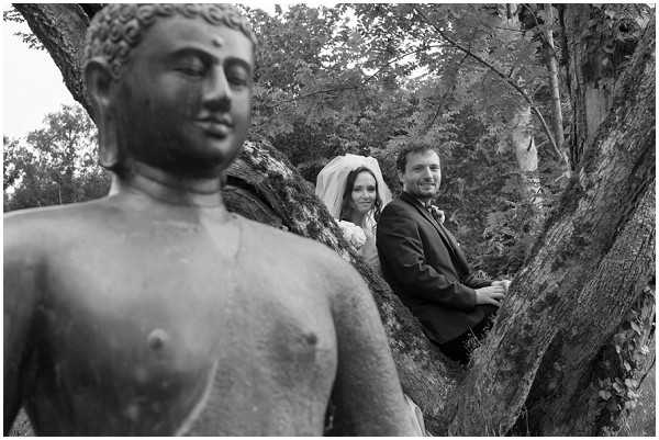 A black-and-white outdoor couple portrait in which the bride and groom are positioned among the branches of a large tree, with the groom in a dark suit and the bride wearing a white dress and veil. In the foreground, a Buddha statue is prominently placed slightly out of focus, creating a layered composition with strong contrast between the statue's smooth surface and the textured tree bark. The shot is taken at a low angle, giving visual weight to the statue while keeping the smiling couple visible in the mid-ground. The overall tonal range is high-contrast with deep blacks in the foliage and mid-grey tones on the statue and couple.