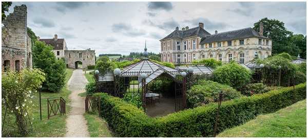 Wide panoramic shot of a French château estate showing the venue grounds on an overcast day. The image captures a large classical French manor house with pale stone facades and multiple windows on the right, connected via formal gardens to older medieval stone ruins and an archway on the left. In the center foreground, an ornate wrought-iron and glass greenhouse or orangerie structure with dark metal framework serves as a focal point, surrounded by neatly trimmed hedgerows and garden pathways. No people or wedding party are visible in this frame. Potential venue feature image.
