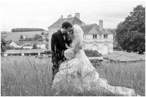 A black-and-white couple portrait taken outdoors in a wild meadow setting, with the bride seated on a large stone and the groom leaning in to kiss her forehead or nose. The bride wears a full ballgown with a long veil and holds a round bouquet of what appear to be garden roses, while the groom is dressed in a dark suit with a bow tie. The image has strong contrast with bright highlights on the dress and soft mid-tones throughout the scene. In the background, a large classical French chateau with ornate facade detailing and a village roofline are visible, establishing the French countryside setting. The composition is a medium-wide portrait shot with the couple positioned slightly left of center.