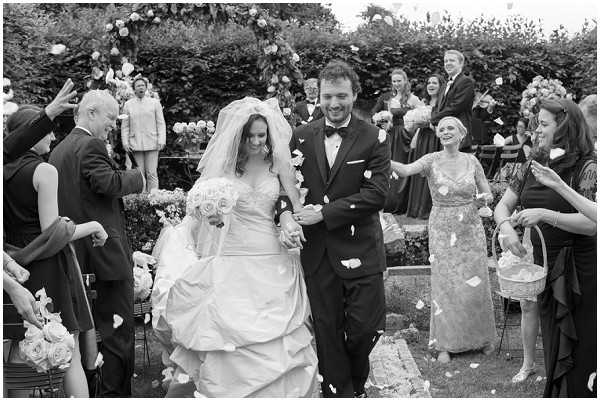 Black-and-white wide shot capturing a bride and groom walking back up the aisle immediately after an outdoor ceremony, surrounded by approximately 20 guests tossing flower petals over the couple. The bride wears a full-skirted ball gown with a cathedral-length veil and carries a large round bouquet of roses, while the groom is dressed in a dark suit with a bow tie and boutonniere. Guests lining the aisle include women in dark knee-length dresses and one in a lace midi dress, several holding small wicker baskets used for the petal toss. The ceremony took place in a formal garden setting, with a floral arch visible at the altar in the background and hedgerow borders framing the space. The image has strong contrast with bright highlights on the bride's dress and veil against the darker-toned crowd.
