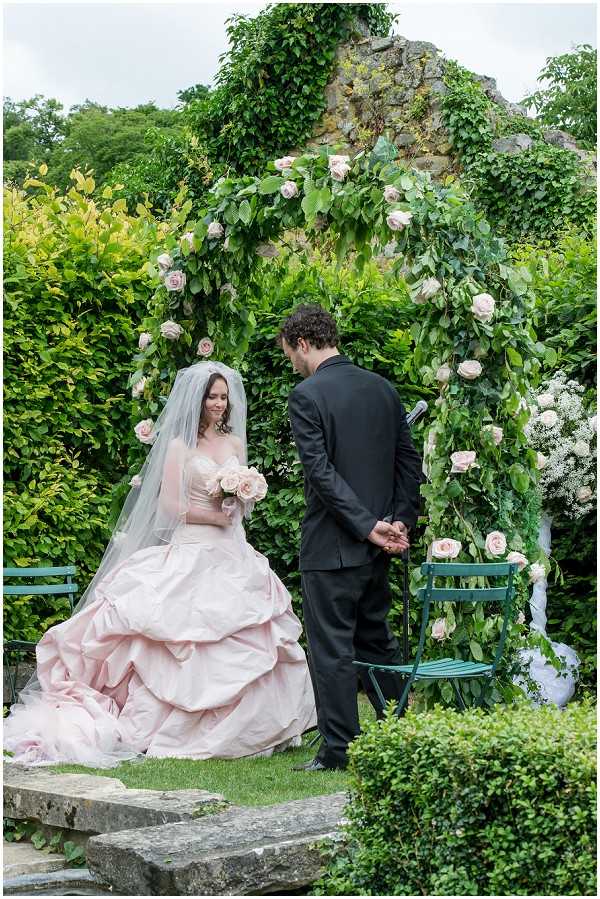 An outdoor garden ceremony captured in a medium portrait shot, showing a bride and groom exchanging vows or rings in front of a large floral arch. The arch is constructed from lush green foliage and blush pink roses, framing a vine-covered stone ruin wall in the background. The bride is seated on stone steps wearing a full-skirted blush pink ball gown with a strapless sweetheart neckline and a cathedral-length veil, holding a bouquet of blush pink roses. The groom stands with his back partially to the camera in a black suit, appearing to hold a ring or read vows. Two teal-painted metal garden chairs are visible near the arch, and the overall decor palette combines blush pink and lush greenery in a romantic garden style.