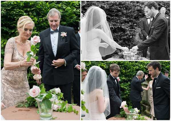 A triptych of images capturing an outdoor wedding ceremony ritual, likely a unity or symbolic rose ceremony. In the largest left image, a woman in a champagne/blush lace long-sleeve dress and a man in a black tuxedo with bow tie and a blush boutonniere stand at a table arranging soft pink roses in glass vases. The top right black-and-white image shows the bride in a strapless gown with a long cathedral veil leaning over a silver tray or vessel, with the groom in a dark suit assisting; the image has soft contrast with mid-range tones. The bottom right color image shows the bride from behind in her veil, facing two men in dark suits and an older gentleman, all gathered around what appears to be the same ceremony table with pink roses, with additional guests visible in the background. The setting is an outdoor garden with dense greenery. All three frames are close-to-medium portrait and detail shots documenting the same ceremonial moment from different angles.