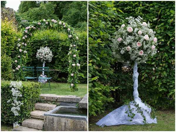 A side-by-side detail shot of outdoor ceremony floral decorations set in a garden. The left image shows a circular floral arch decorated with blush pink roses, white gypsophila, and lush greenery, framing stone steps leading to a small water feature, with teal garden chairs visible in the background. The right image shows a tall topiary-style floral column featuring a ball-shaped arrangement of blush pink roses and white gypsophila on a draped white fabric-wrapped stem, with trailing ivy at the base. Both arrangements share a consistent blush and white floral palette in a classic garden style.
