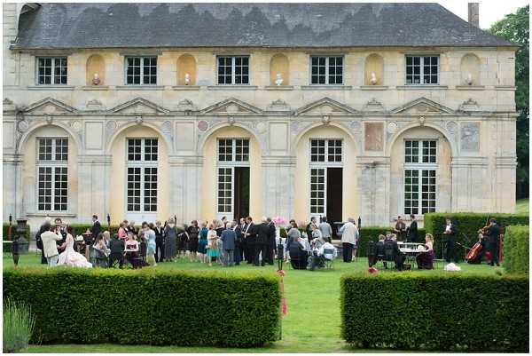 A cocktail hour is taking place outdoors on the manicured lawn in front of a large French classical chateau or orangerie with pale stone façade, arched windows, and ornamental reliefs. Approximately 40-50 guests are mingling across the grounds, with the bride in a white gown visible on the left side of the group. A string ensemble of several musicians with chairs and music stands is set up to the right of the gathering. The scene is framed by neatly trimmed formal hedgerows in the foreground, and the overall styling appears classic and formal. Wide shot taken from a distance capturing both the architectural backdrop and the full scope of the outdoor reception. Potential venue feature image.